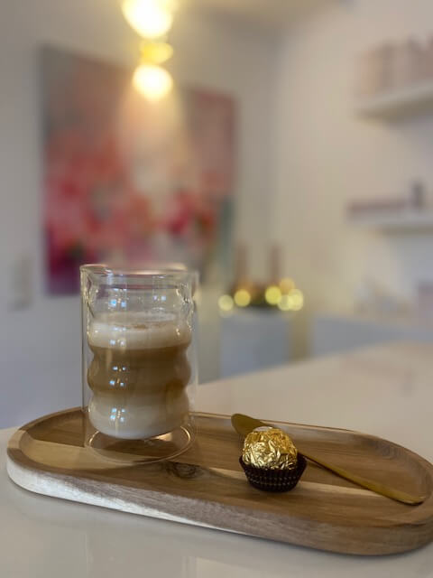 Ein Latte Macchiato und ein Pralinenkonfekt auf einem Holztablett schaffen eine entspannte Atmosphäre im Kosmetikstudio.