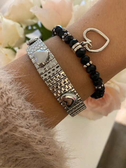 Zwei elegante Armbänder mit Herzmotiven schmücken ein Handgelenk, während im Hintergrund zarte Blüten Kosmetikstudio-Flair verbreiten.