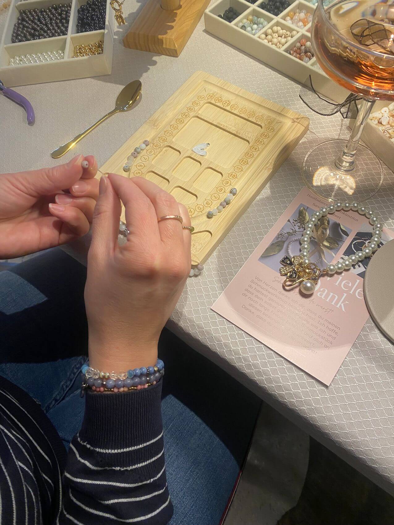 Eine Person im Kosmetikstudio erstellt mit Perlen ein individuelles Schmuckstück, während ein Glas Wein daneben steht.