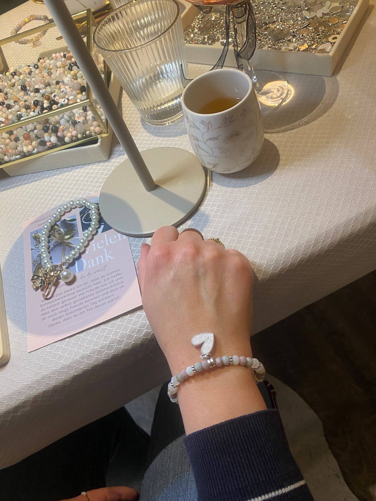 Eine Hand mit einem eleganten Armband liegt auf einem Tisch, umgeben von Kosmetikprodukten und Schmuck.